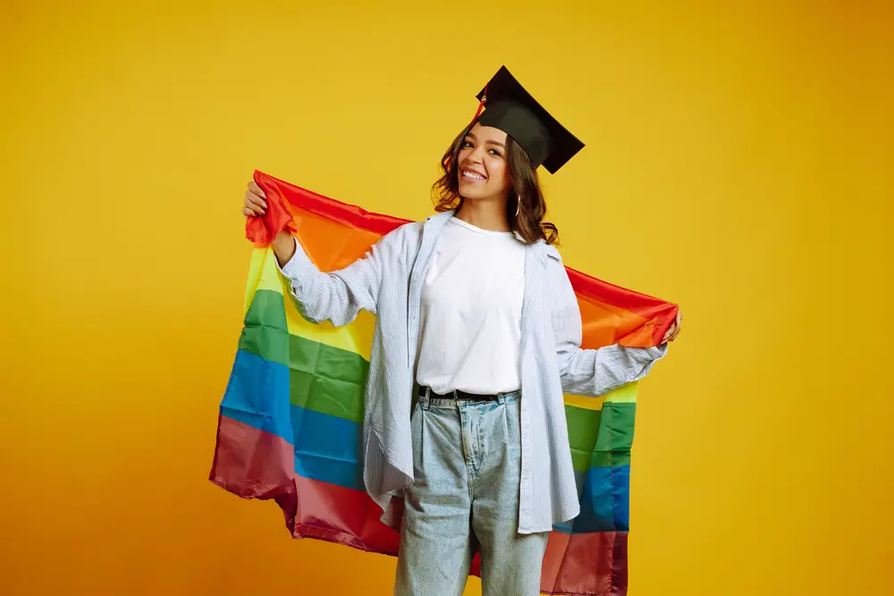 smiling-african-american-woman-in-graduation-hat-posing-with-rainbow-flag-against-yellow-backdrop