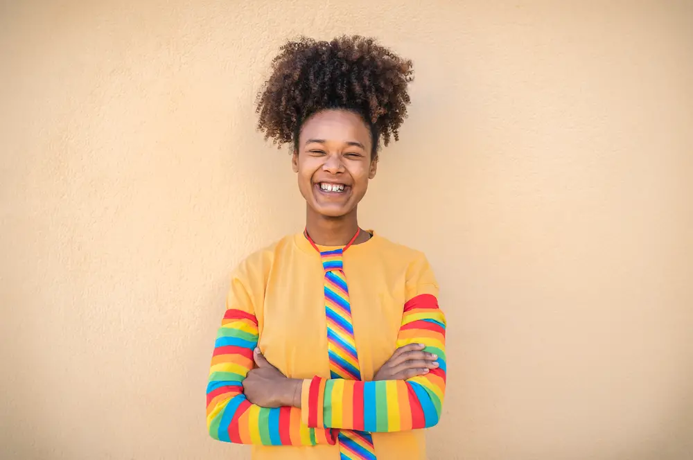smiling-afro-woman-in-rainbow-colored-tee-shirt-standing-with-arms-folded-against-light-brown-backdrop