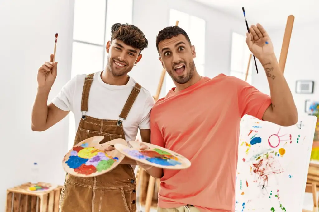 two-hispanic-men-smiling-at-camera-holding-paintbrushes-and-palette-in-painting-class