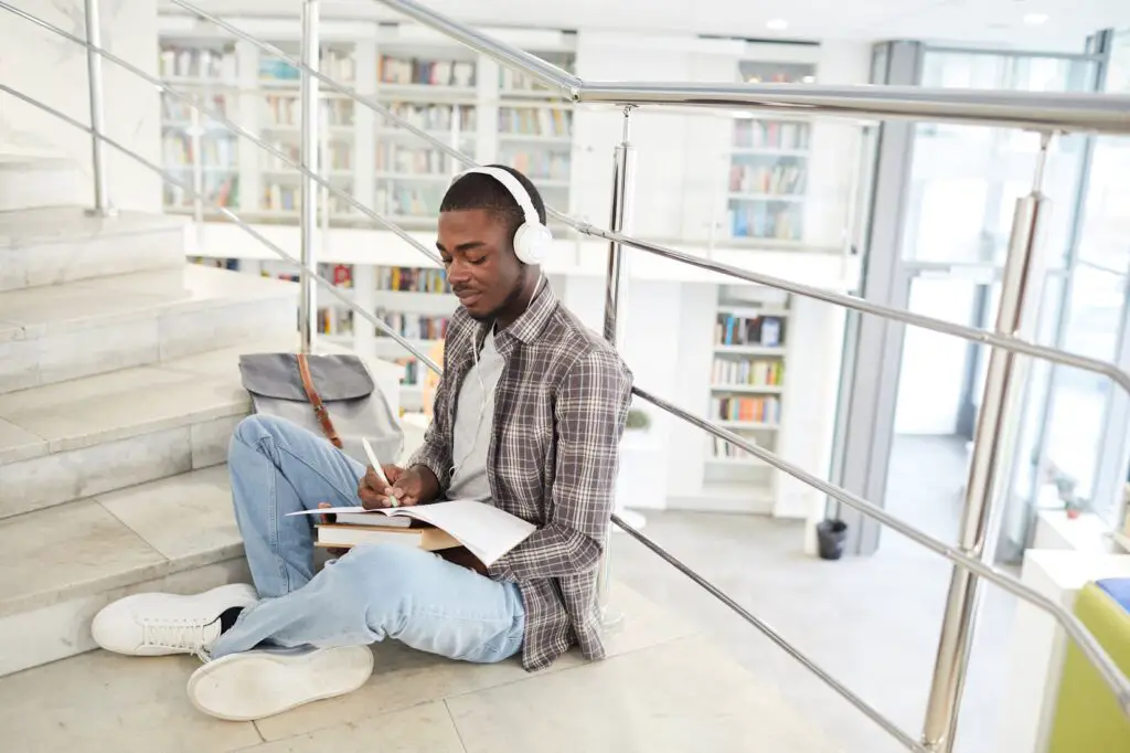 a-male-college-student-with-white-headphones-dividing-his-study-materials-sitting-by-the-stairs-in-a-library