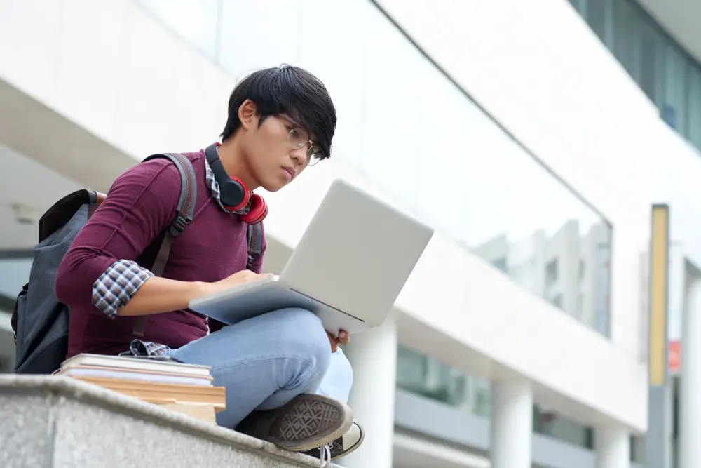 A seated Asian male high school senior focuses on preparing his early decision college application documents on his laptop