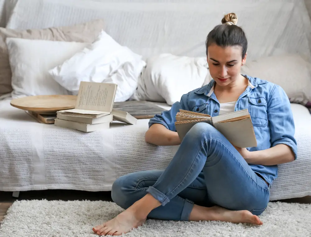 A female sophomore reads one of the best books for college students on her bedroom floor beside her bed stacked with four other books