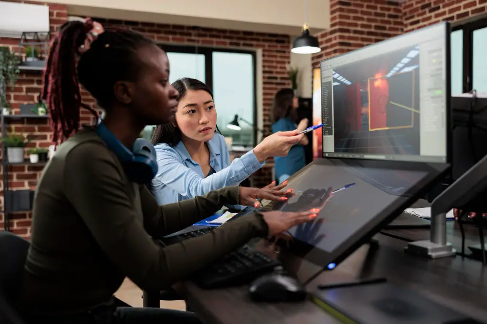 An Asian female mentor guides an African-American female game design student on a computer and tablet