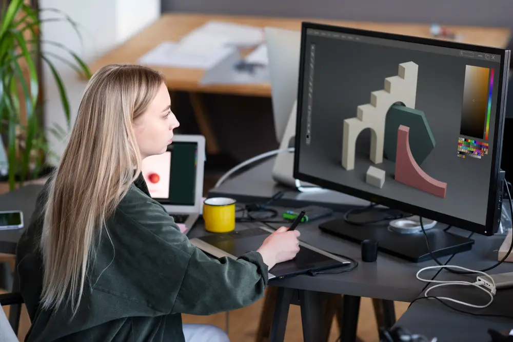 A Caucasian female game design student draws 3D forms on her tablet while facing the desktop screen