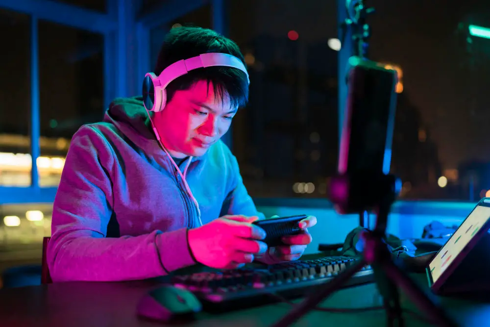 An Asian male college student focuses on playing a mobile game at night after studying