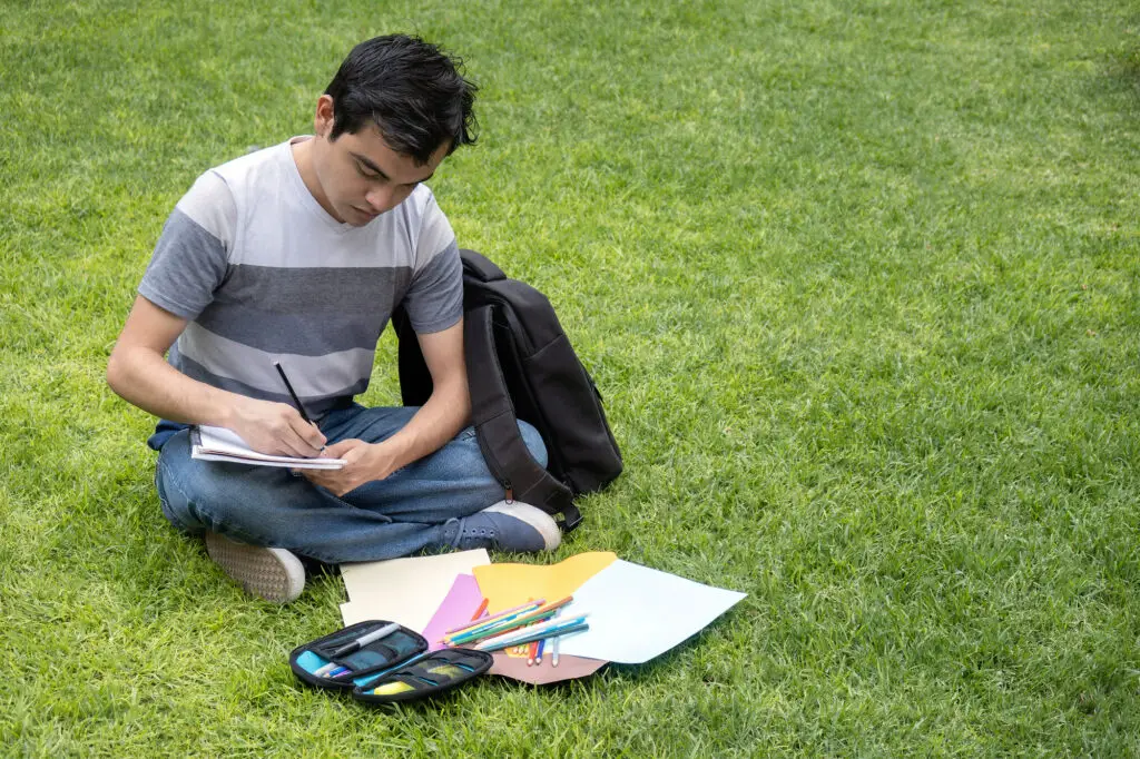 A young man sitting on the grass using a notebook, markers, and papers as college study hacks