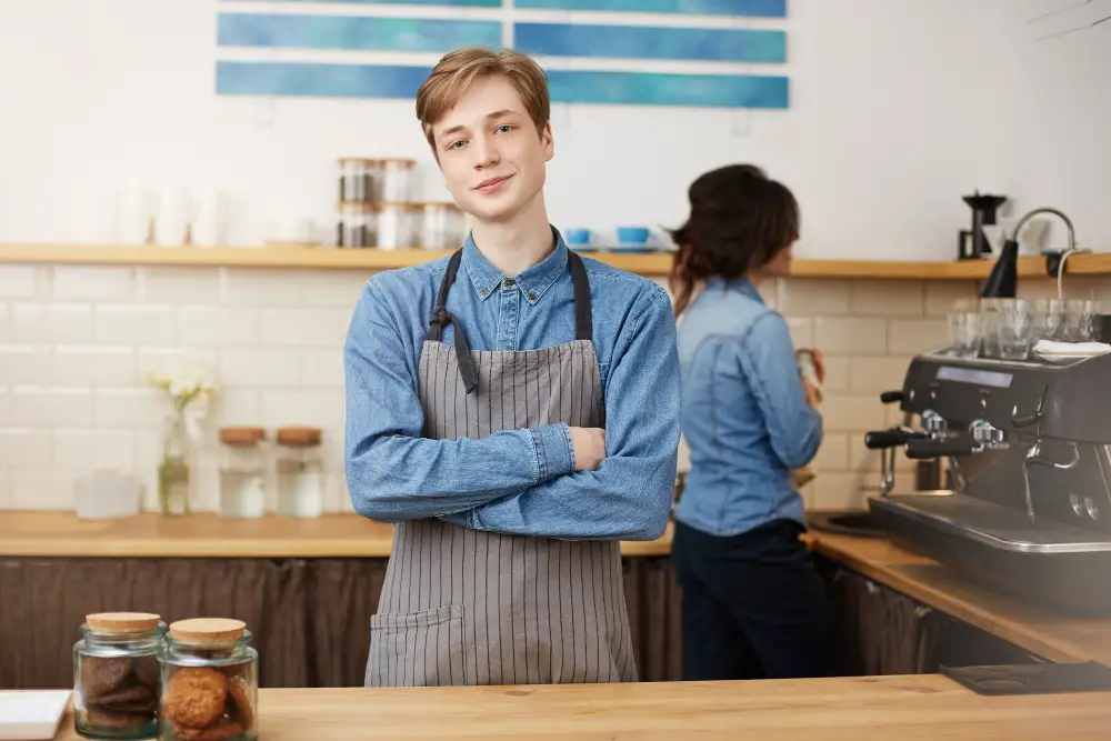 A blond male student smiling while standing behind the counter of one of the fast food jobs that pay for college