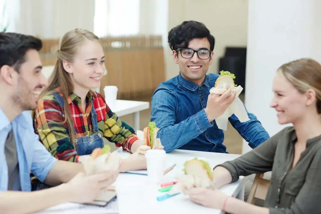 Four young people enjoy sandwiches together at a table, sharing a meal, and engaging in conversation