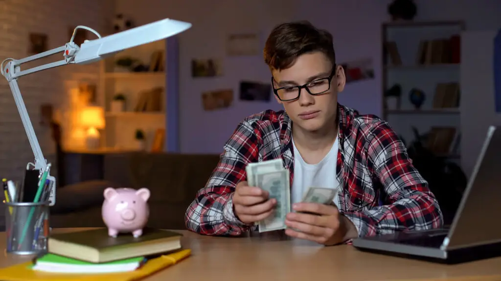A young man sits at a desk with money and a piggy bank, managing his finances responsibly before deciding about campus housing.
