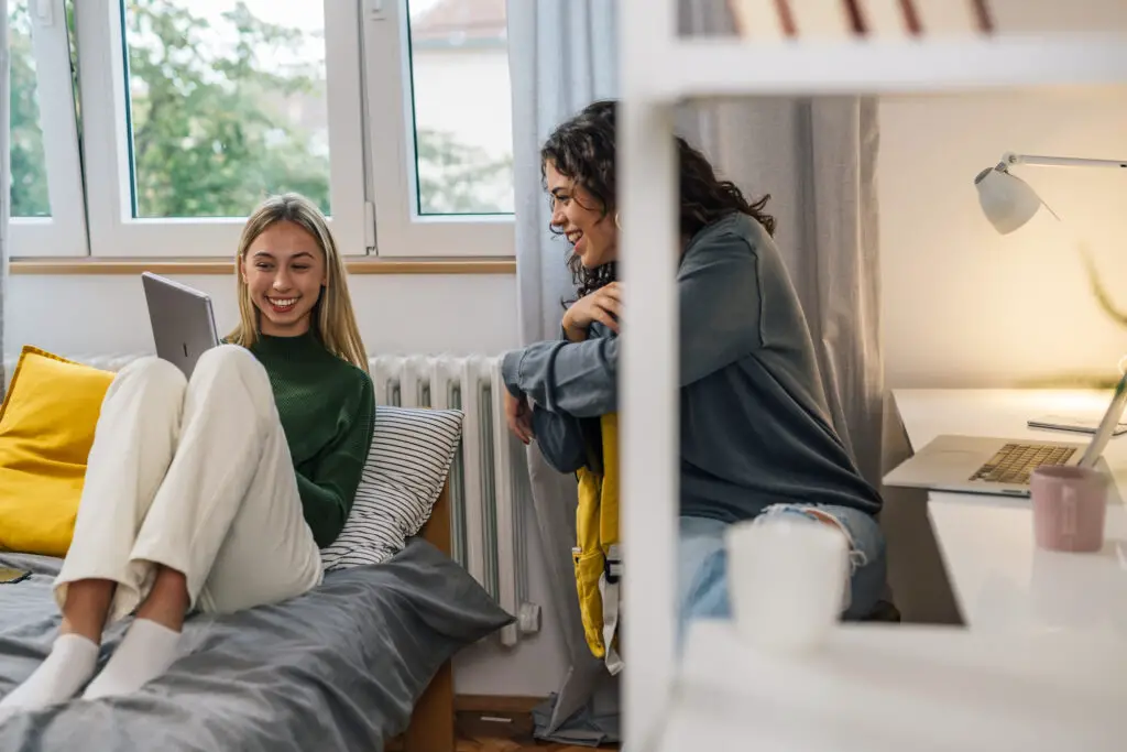 Two college women sitting on a bed inside their campus housing, engrossed in their laptop work.