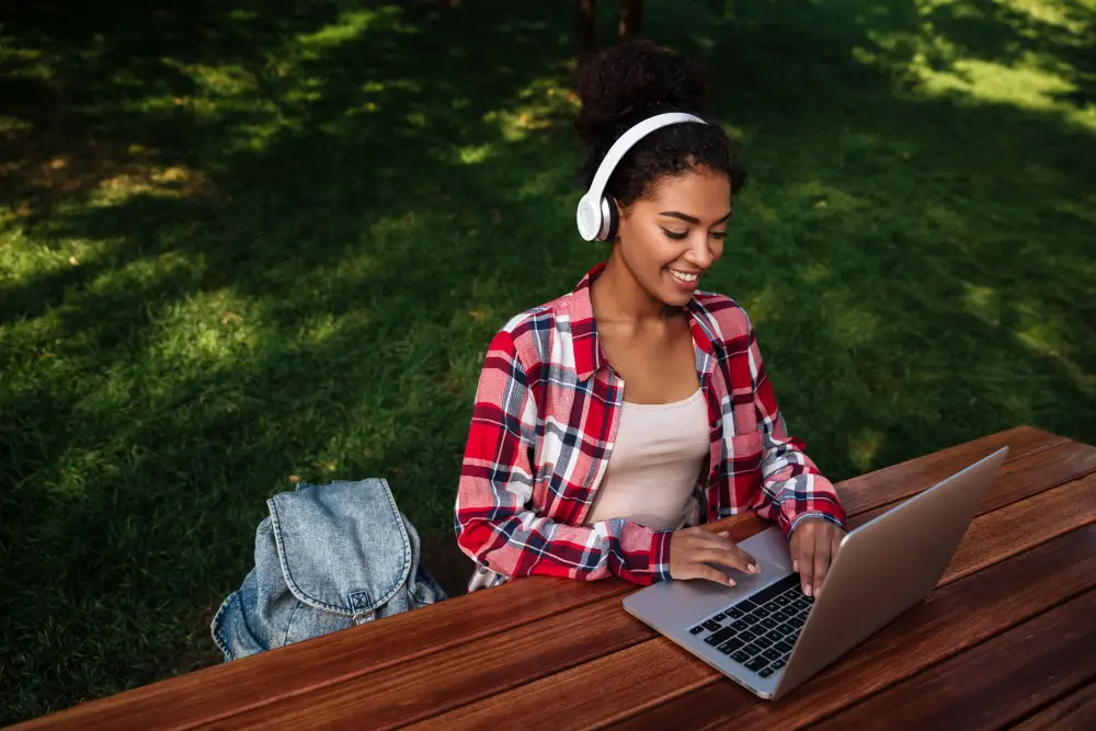 A smiling female college student wearing headphones while checking notes in her laptop outdoors