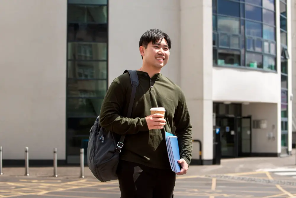 A smiling male Asian college student carrying his backpack, reusable coffee cup, and folders on the way to class