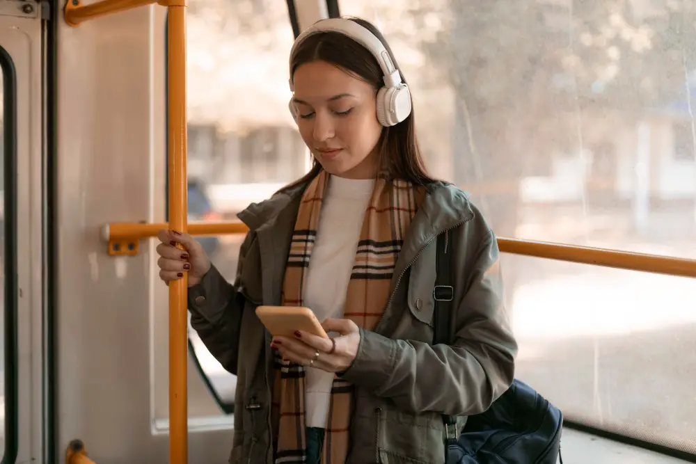 A female college student wearing headphones checks her phone while riding the bus to university