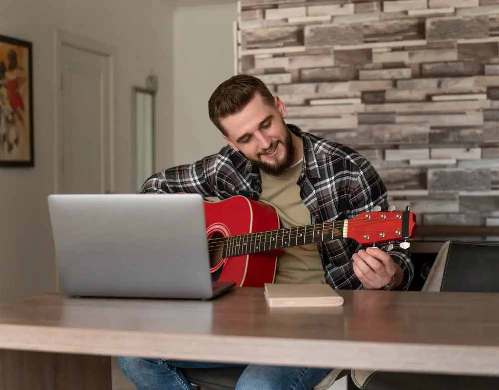 A smiling male college student tunes his guitar based on online video tutorials