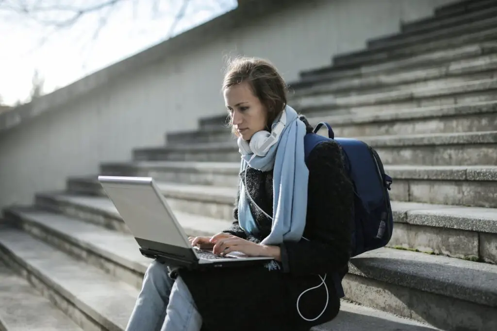 A young woman wearing a backpack while sitting on steps with a laptop, working outdoors