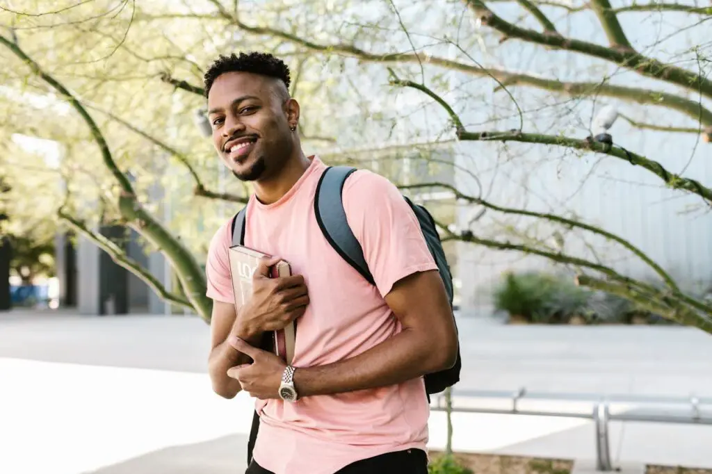 A smiling young man wearing a timeless watch, a pink shirt, and a backpack