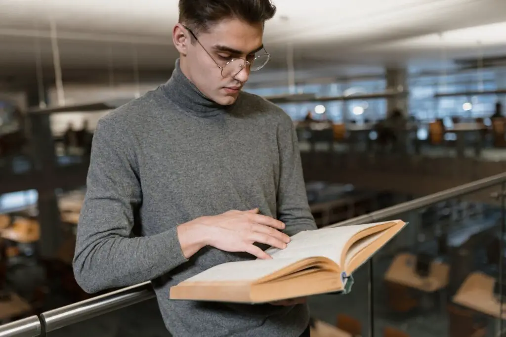 A man with glasses and cozy sweater while reading a book in a library