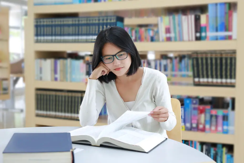 A woman with glasses reading a book in a library to prepare for BAR exam