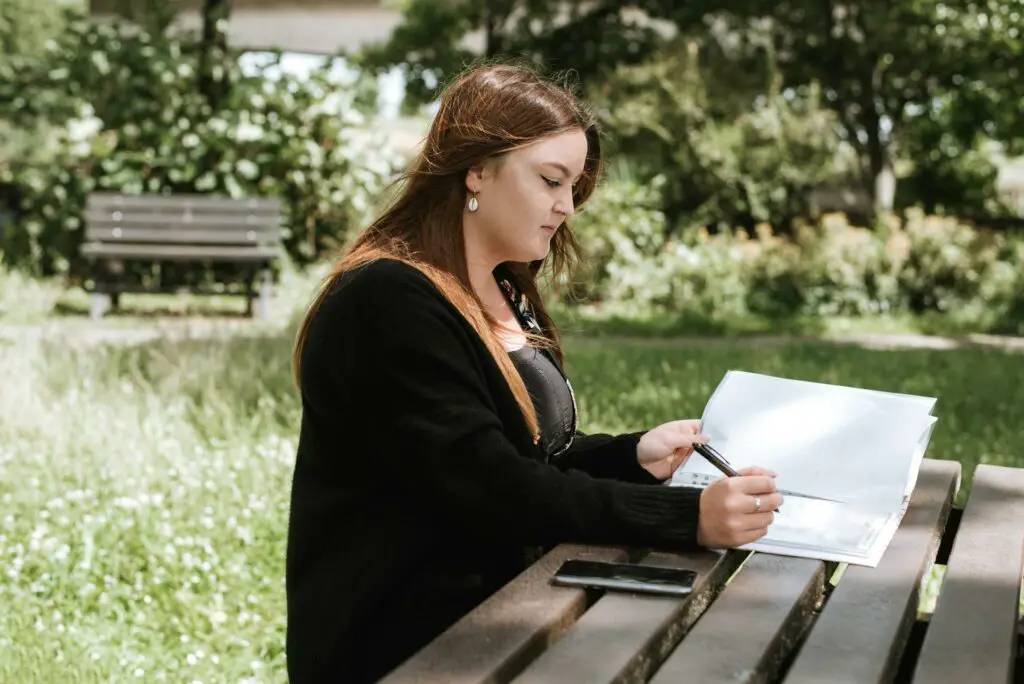 A woman sitting on a bench outdoors focused on reading a piece of paper.