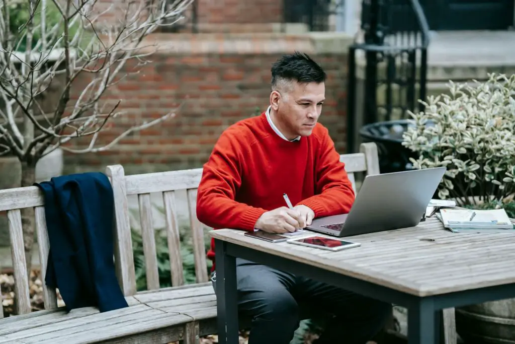 A man in a red sweater is sitting on a bench, using a laptop.