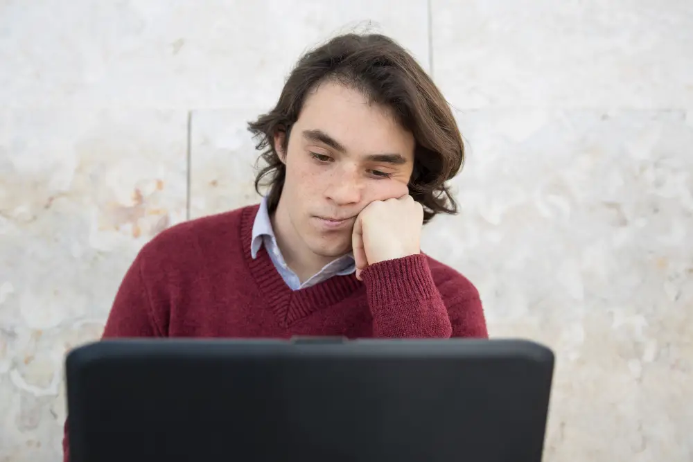 A male high school senior frowns in disappointment after reading a college rejection letter sent via email on his laptop