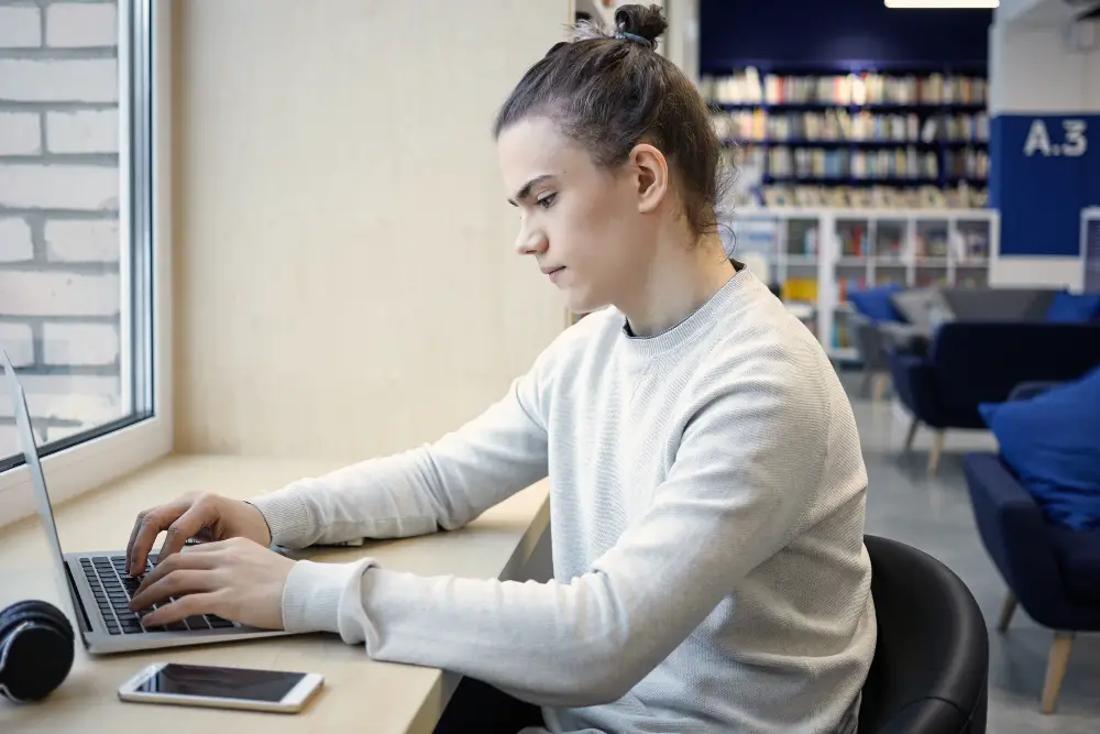 A focused blond male college student writing a paper on his laptop beside his phone in the college library