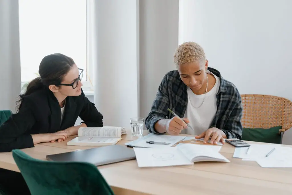 A college student and professor are seated at a table, engrossed in their work, surrounded by papers and a pen