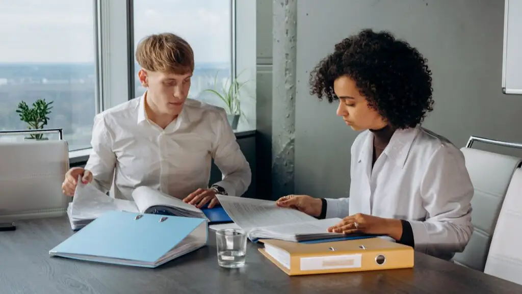 Two college students seated at a table, engaged with papers and a laptop, collaborating on a project or discussion to avoid plagiarism