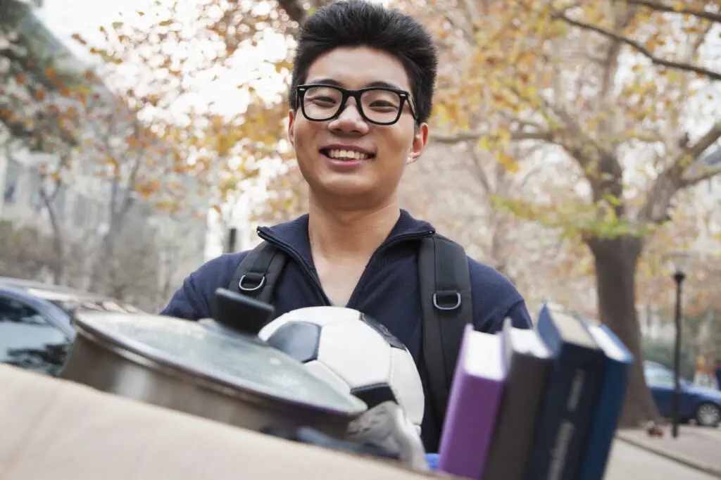 A young student with glasses and a backpack holds a box with a soccer ball, books, and other dorm essentials