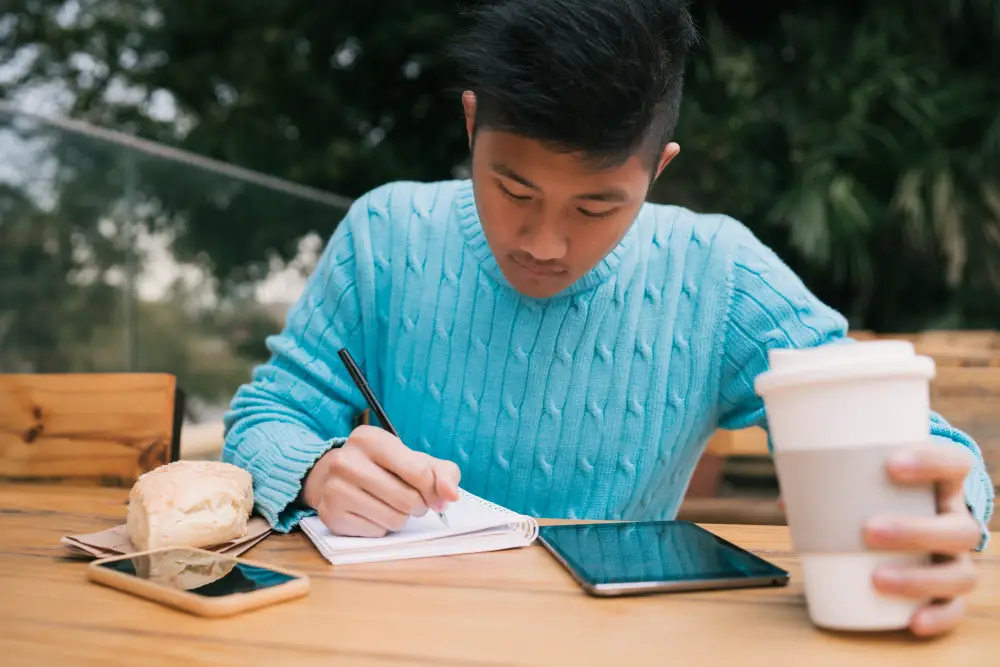 A male gap year program participant writes about his activities and reflections on his journal while holding a cup of coffee